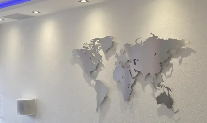 Weltkarte aus Edelstahl in einem Wohnzimmer hängend. Vor einer weissen Wand. Unterhalb ist ein Sofa zu sehen.